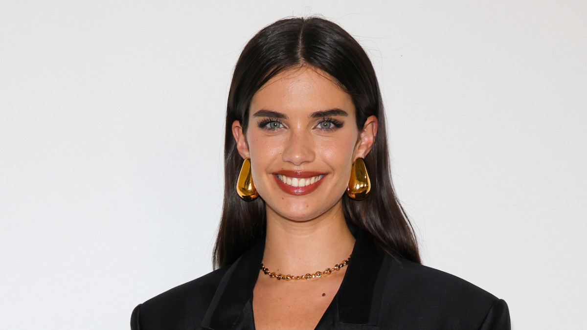 Sara Sampaio poses in front of a white backdrop in a black blazer, red lip and oversized gold earrings.